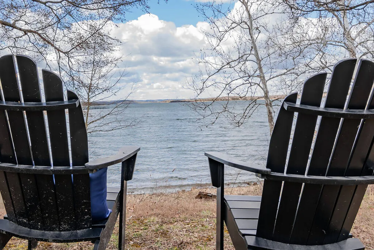 Two black Adirondack chairs overlook the calm lake beneath a cloudy sky, set just steps from the shoreline. The setting exudes tranquility and invites quiet relaxation, reading, or sharing a glass of wine while enjoying the sunset or stargazing at night.