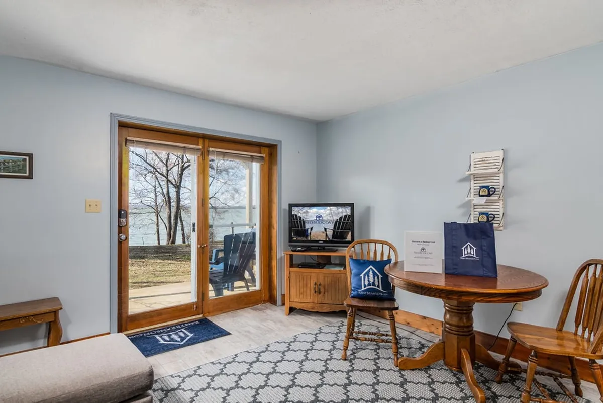 A spacious interior scene showcasing large sliding glass doors that frame a stunning outdoor view, a round wooden dining table for two with cushioned chairs, and a television for entertainment. The room is filled with soft, natural light, creating a cozy, inviting setting that encourages togetherness and relaxation.