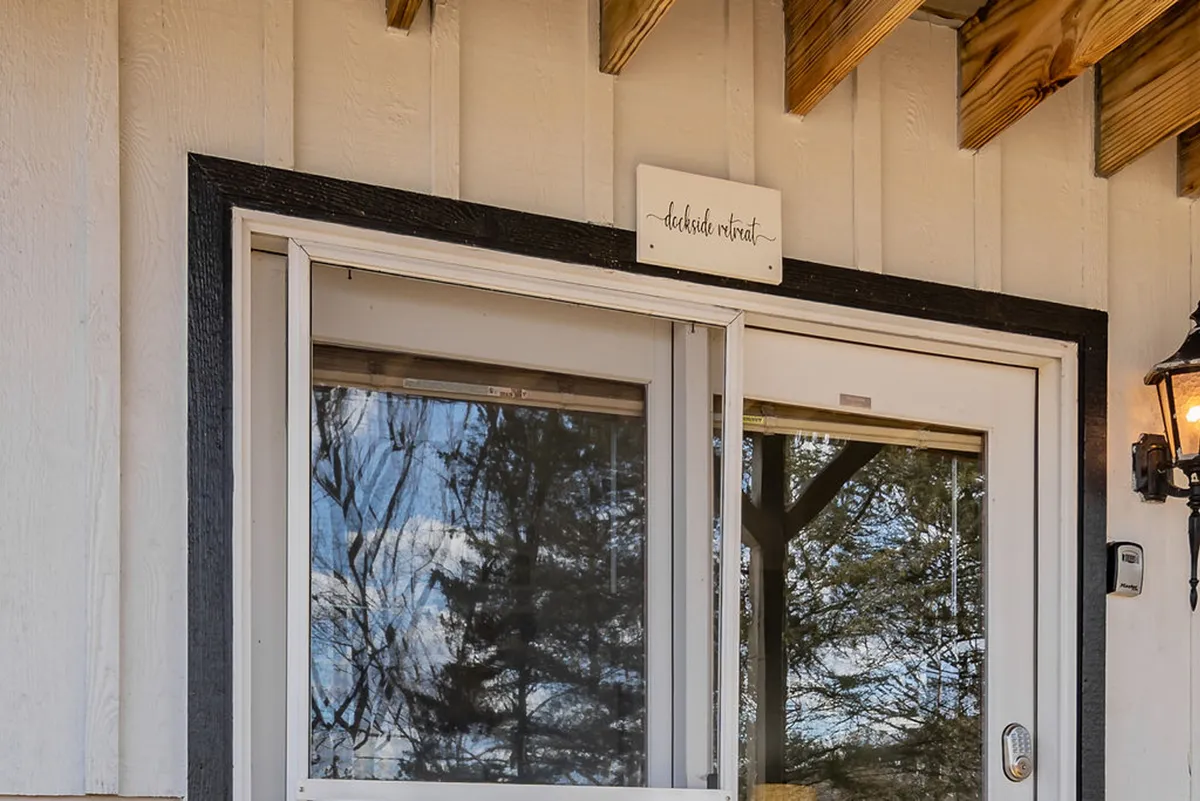 An inviting view of the entryway to Dockside Retreat, highlighting the doorway, a sign reading 'deckside retreat,' and reflective windows. The surrounding deck and visible greenery suggest a welcoming environment with seamless flow between indoor comforts and outdoor relaxation.