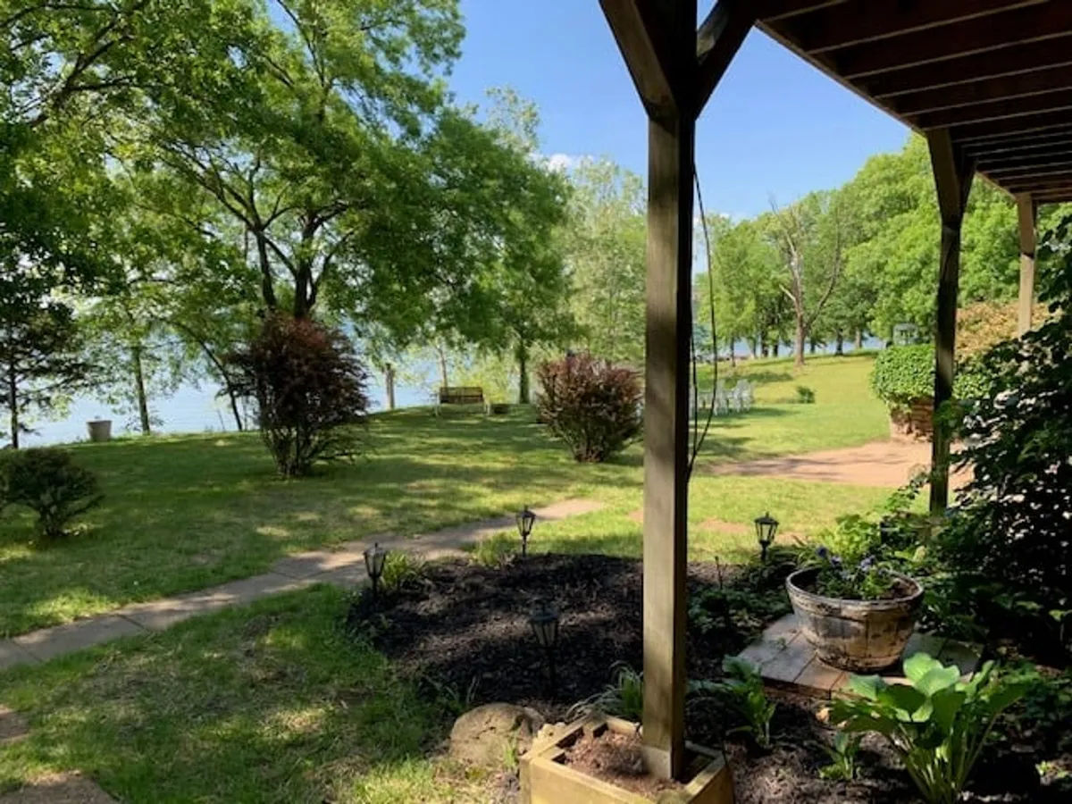 A peaceful lakeside scene with lush green trees framing a serene waterfront view. A garden path leads toward the water, inviting guests to stroll from their private patio down to the lake's edge. The soft sunlight hints at the possibility of both tranquil mornings and romantic sunset moments by the lake.