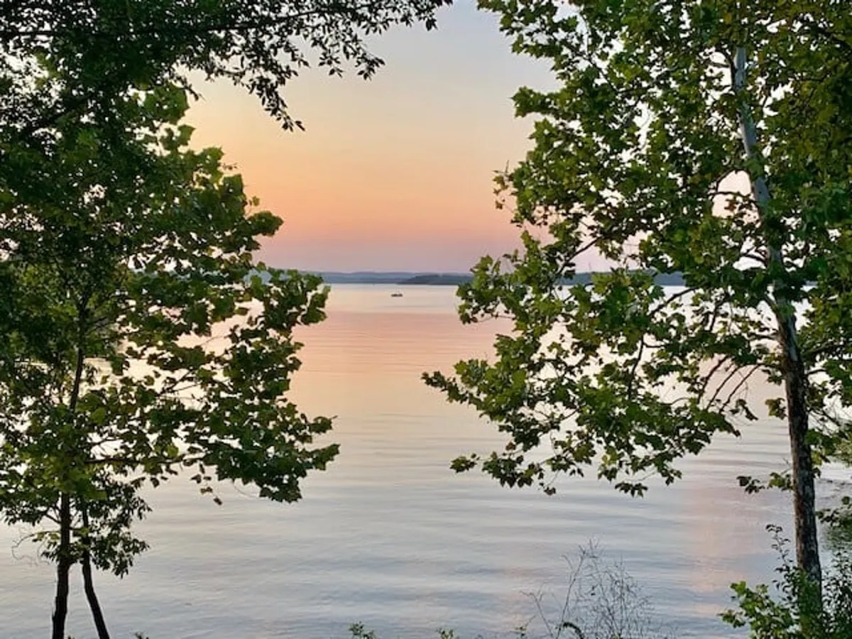 A wide-angle view of the lake framed by trees, with the sun setting in the background and reflections shimmering on the water. The tranquil scene highlights the beauty of sunset over Table Rock Lake, inviting thoughts of evening relaxation after a day of adventure.