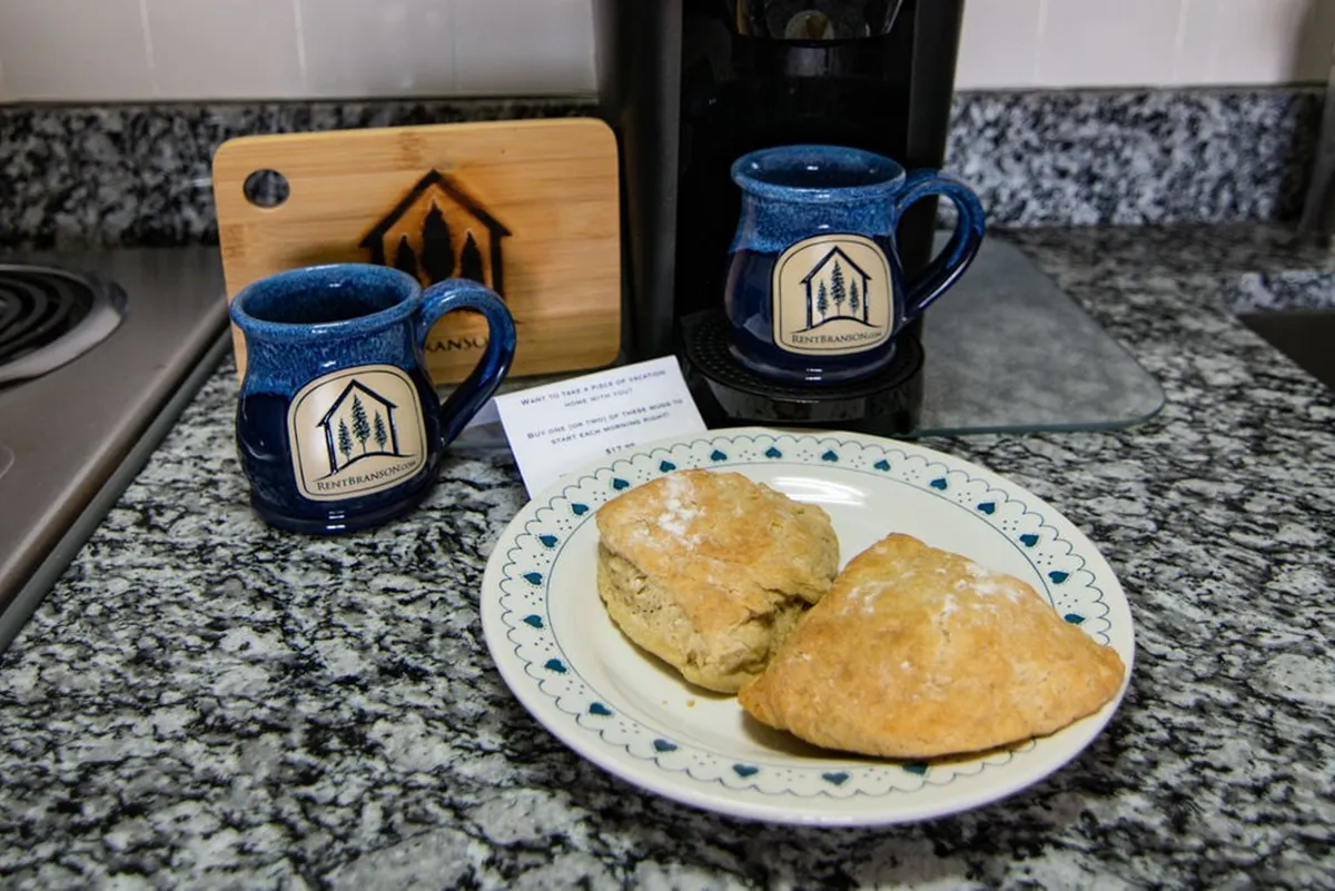 A close-up of the dining experience, showing two blue mugs and a plate with scones on a wooden table, accompanied by a coffee maker and cutting board in the background. The image exudes a cozy, welcoming atmosphere ideal for relaxed breakfasts or casual get-togethers.