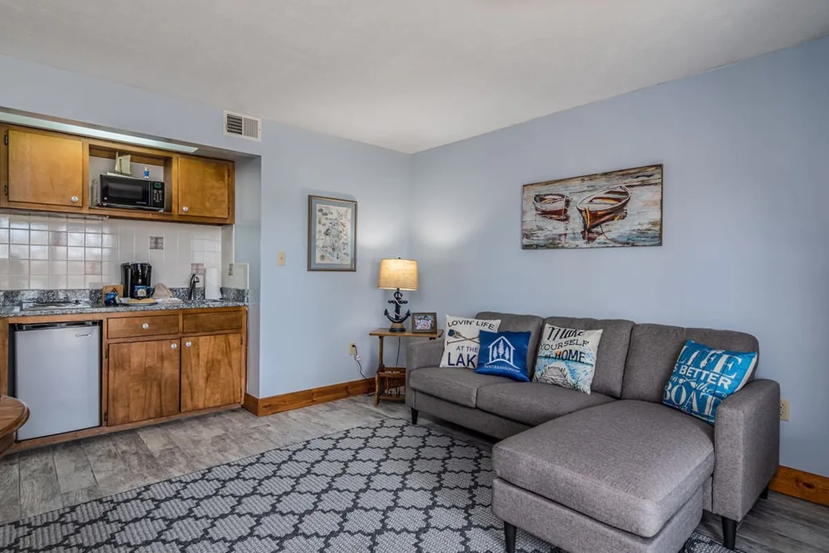 A wide angle of the suite’s living area, featuring a comfortable sofa, kitchenette in the background, a lamp, and wall artwork. The space feels lived-in and intimate, showcasing the relationship between cooking, dining, and relaxing.
