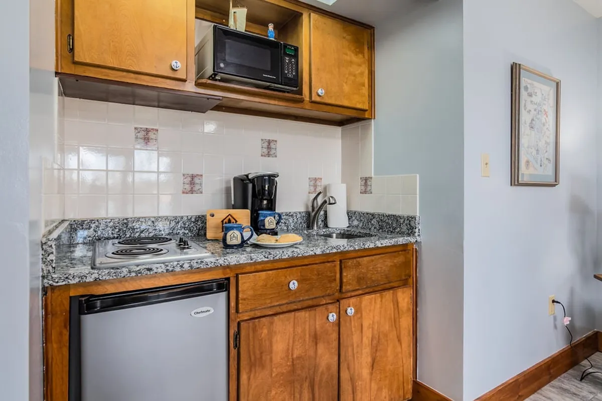 A practical kitchenette featuring a granite countertop, compact coffee maker, microwave, kitchen cabinets, and a small refrigerator. The setup is streamlined for self-catering and features all essentials for meal prep and morning routines.