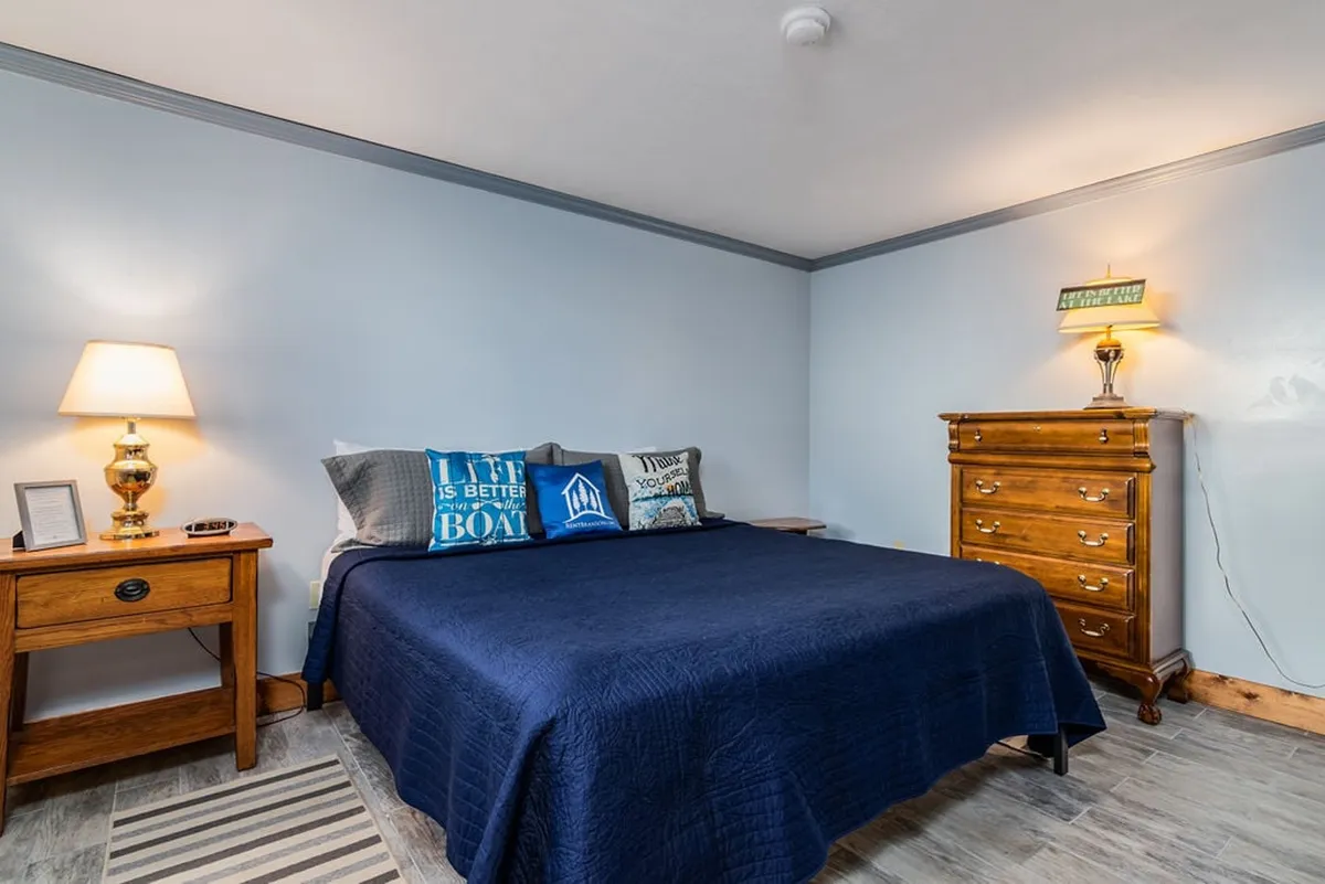 A serene bedroom featuring a king-size bed dressed in blue bedding, accompanied by decorative pillows and wooden nightstands with lamps. The space exudes a calm and restful atmosphere, ideal for unwinding after a day by the lake.