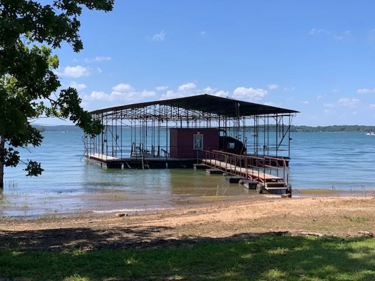 The image displays a classic waterfront dock next to a boathouse structure, with trees providing shade. The dock serves as a hub for fishing, swimming, boating, and easy kayak or paddleboard launching, with the tranquil lake inviting adventure and relaxation.