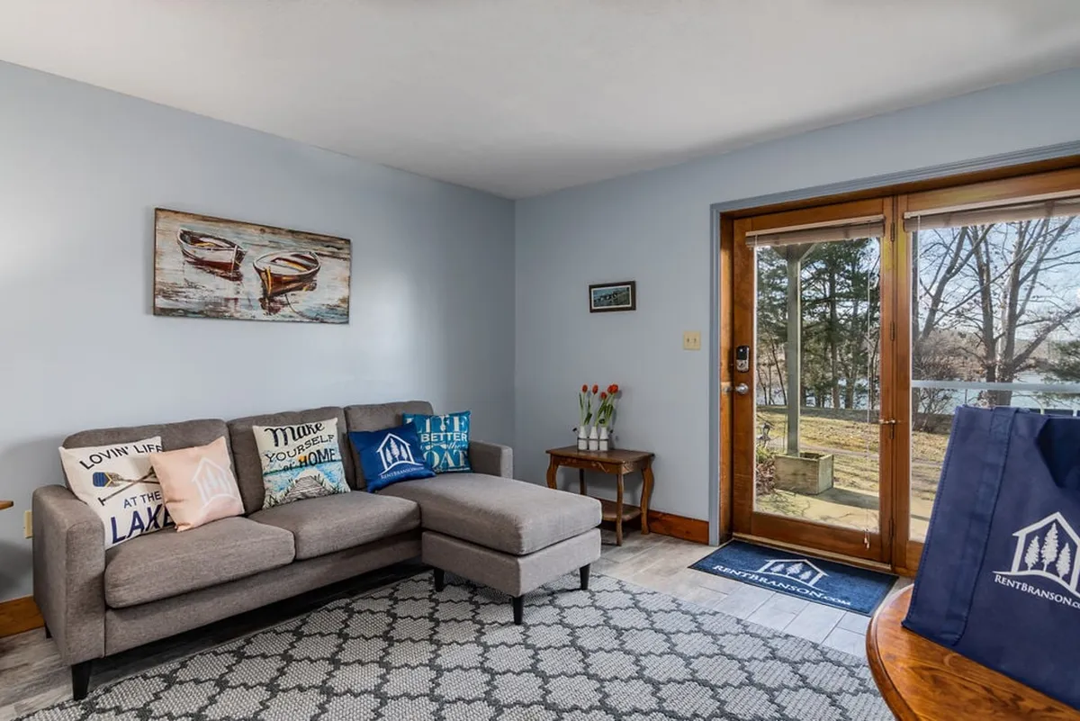 This image features a cozy gray sofa with decorative pillows, positioned to face a large window and a wooden door that leads directly to an outdoor area. A small table with flowers and artwork on the wall contribute to a welcoming, homelike atmosphere. Through the window, you can glimpse natural light that hints at scenic lake or garden views.
