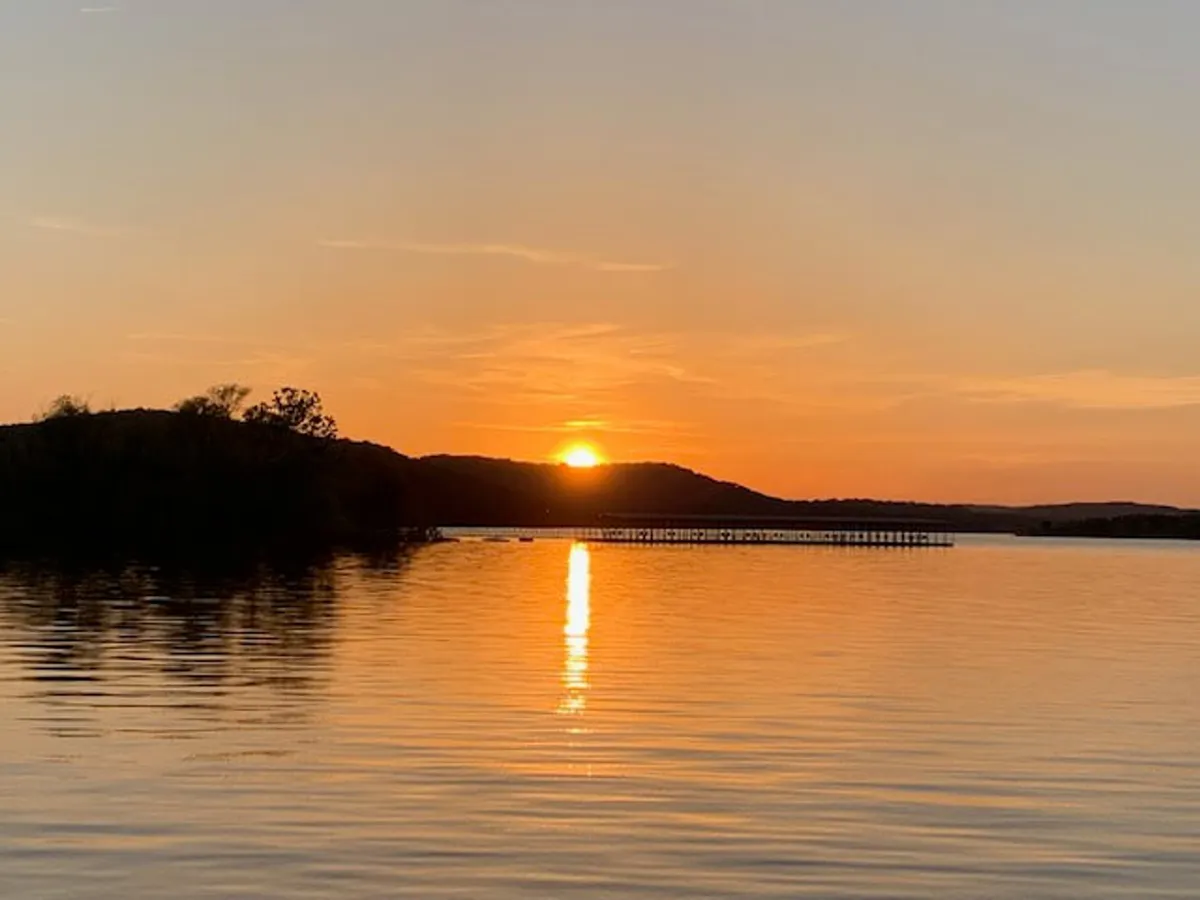 The image captures a tranquil lakeside scene at sunset, with calm waters reflecting the colorful sky. A pier extends toward the horizon, framed by silhouetted hills, evoking a sense of peace and connection with nature. The setting invites activities like fishing, boating, and quiet reflection.